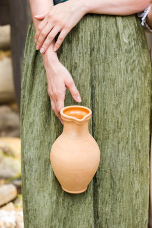 Closeup of Hands of Caucasian Girl Holding Clay Jar in Front of Decorated Dress Outdoor. Vertical Shotの写真素材