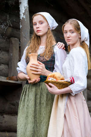 Rural Style Concepts. Two Young Beautful Caucasian Girlfriends in Traditional Rural Outfit  Posing With Caly Jar And Bread Together Outdoor. Vertical Shotの写真素材