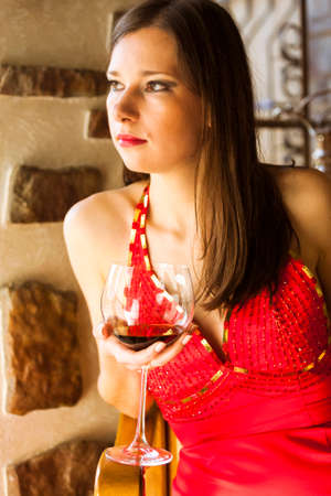 Dreaming Caucasian Brunette Female in Long Red Dress Posing with Large Glass of Wine Against Big Window on Background Indoors. Vertical imageの写真素材
