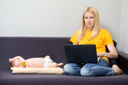 Distant Working Concepts. Young Caucasian Blond Mother Working With Laptop On Couch in Front of Her Newborn Male Child.  Horizontal Imageの写真素材