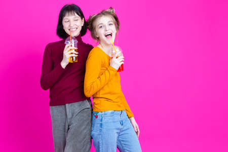 Two Laughing Smiling Caucasian Girls Together with Cups Drinking Colorful Juice or Smoothie With Straw Over Pink Background. Horizontal Imageの写真素材