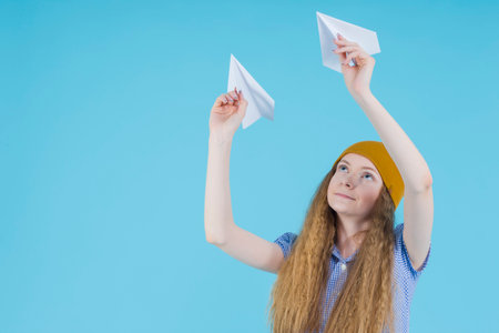 Blond Teenager Girl in Blue Checked Dress,  Warm Hat and Rubber Boots Launching Pair of Origami Paper Planes Over Blue Background. Horizontal Image Orientationの写真素材
