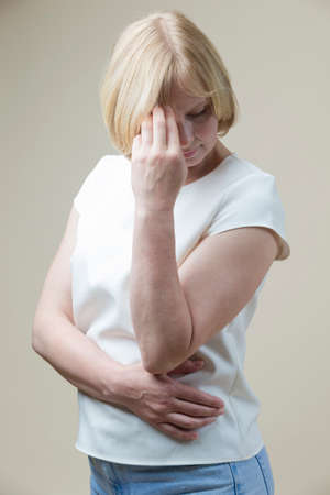 Depressed and Sad Caucasian Mature Woman Suffering From Headache With Lifted Hands Against Beige....