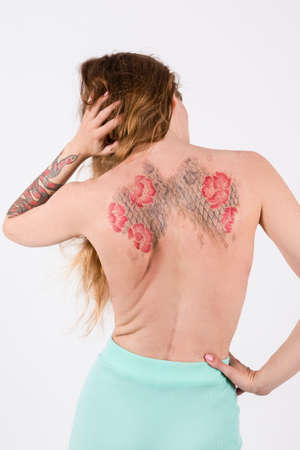 Back View of Young Caucasian Brunette Woman With Flowery Tattoes on Her Back Posing With Lifted Hand On White. Vertical Shotの写真素材