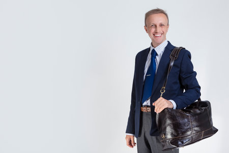 Young Positive Caucasian Man in Blue Suit Holding Brown Leather Bag Over Shoulder and Posing Against White. Horizontal Image Compositionの写真素材