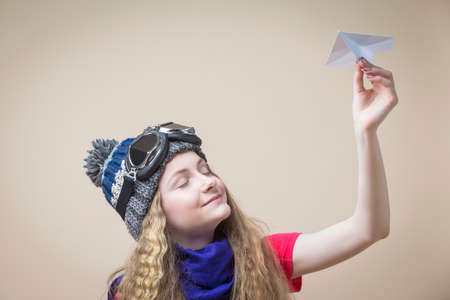 Dreaming Teenage Girl in Flying Glasses And Knitted Warm Hat With Origami Paper Plane With Closed Eyes Over Beige Background. Horizontal Imageの写真素材