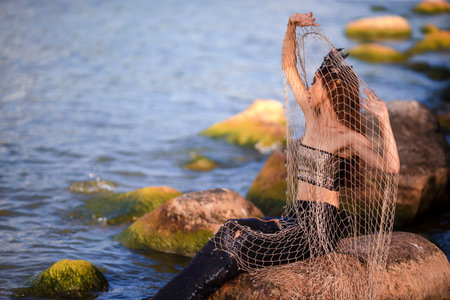 Sensual Mermaid With Net At Rocky Sea Coast Wearing Seashell Decorated Crown and Black Shiny Tail On Slim Body Covered With Strasses Posing As Mistress Queen of Sea. Horizontal Image Compositionの写真素材
