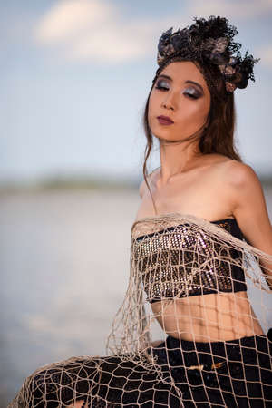 Mermaid With Net At Rocky Sea Coast Wearing Seashell Decorated Crown Posing As Mistress Queen of Sea. Vertical Shotの写真素材