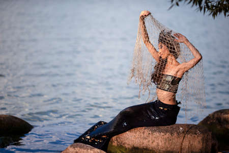 Asian Mermaid With Net At Sea Coast on Rocks While Wearing Seashell Decorated Crown and Black Shiny Tail On Sexy Body Covered With Strasses As Mistress Queen of Sea. Vertical Shotの写真素材
