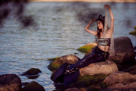 Asian Mermaid With Net At Sea Coast on Rocks While Wearing Seashell Decorated Crown and Black Shiny Tail On Sexy Body Covered With Strasses As Mistress Queen of Sea. Vertical Shotの写真素材