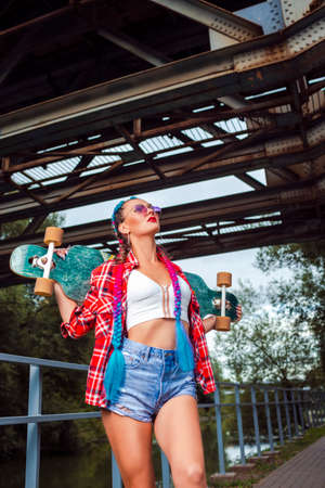 Hipster Skateboarder Girl Posing With Longboard Under Rusty Metal Construction Outdoor While Holding  Skateboard Behind and  Wearing Sunglasses. Vertical Imageの写真素材