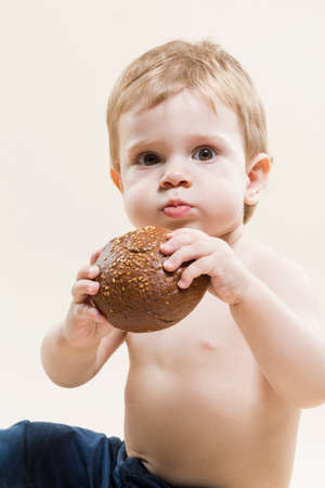 Portrait of Little Child With Food in Hands While Eating Freshly Baked Bun And Posing Against Beige...
