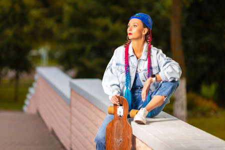 Lifestyle Concepts. Bright Caucasian Female With Longboard Sitting on Brick Fence in Cap While...