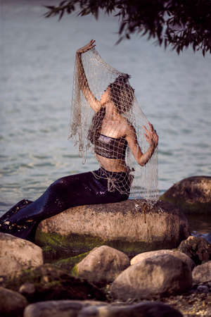 Mermaid Caught In Net At Sea Coast on Rocks Wearing Seashell Decorated Crown and Black Shiny Tail On Body Covered With Strasses As Mistress Queen of Sea. Vertical Shotの写真素材