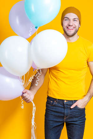 Funny Smiling Caucasian Young Handsome Man With Colorful Air Balloons Posing in Warm Hat Over Yellow Background.Vertical imageの写真素材
