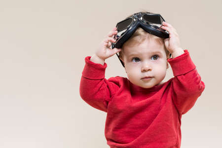 Little Caucasian Child Posing With Aviator Goggles While Sitting Against Beige Background....