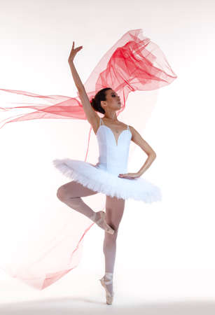 Ballet Ideas. Professional Japanese Female Ballet Dancer Posing in White Tutu With Flying Red Cloth In Hands Against White Background. Vertical Shotの写真素材