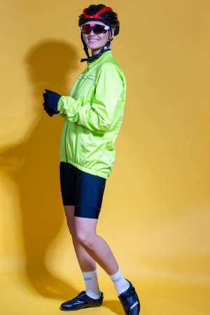 Full Length Portrait of Confident Positive Female Road Cyclist in Professional Outfit Standing In Green Autumn Jacket Against Yellow Background. Vertical Imageの写真素材