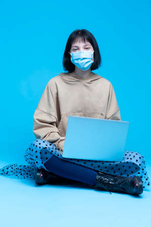 Smiling Caucasian Brunette Girl Sitting with Laptop While Wearing Facial Protective Mask Against Viruses in Hoodie Against Blue Background. Vertical Shotの写真素材
