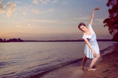 Passion To Dance With Japanese Ballet Dancer in White Dress And Silver Crown Standing In Ocean Wave And Showing Ballet Pas With Lifted Hands Against Picturesque Sunset At Sea.Horizontal Image Compositionの写真素材