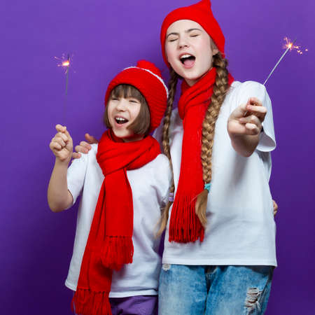 Two Expressive Laughing Caucasian Sisters Girls in Red Warm Hats and Red Scarves Having Fun With Vivid Bright Burning Bengal Lights Fireworks Over Purple Background. Square Imageの写真素材