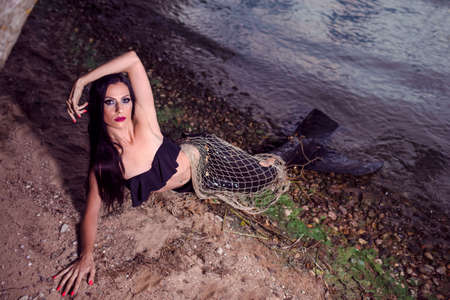 Mermaid Posing With Net Near Sea on Rocks While Wearing Artistic Makeup and Black Shiny Tail Covered...