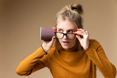 Young Caucasian Blond Girl Listening To Arcane Hidden Gossips and Secrets Using Disposable paper Drinking Cup Over Beige. Horizontal imageの写真素材
