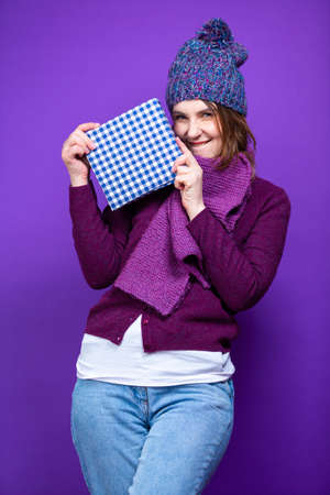 Caucasian Female Woman in Warm Knitted Hat and Violet Scarf Posing with Big Checked Wrapped Present Gift Box And Smiling Happily Over Purple  Background.Vertical Imageの写真素材