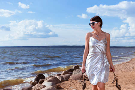 Portrait of Sensual Caucasian Sexy Brunette Female Girl in Light Dress Holding Shoes While having rest and Relaxing On Shoreline Outdoors At Sunny Day. Vertical imageの写真素材