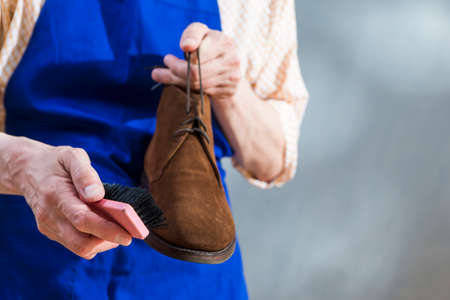 Professional Male Shoes Cleaner In Apron Holding Brown Suede Chukka High Boot With Brush Against Gray Background in Workshop. Horizontal Imageの写真素材