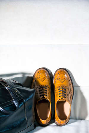 Pair of Male Tanned Full Broggued Oxford Calf Leather Shoes Along With Dark Brown Leather Business Bag on Pale Sofa. Vertical Shotの写真素材