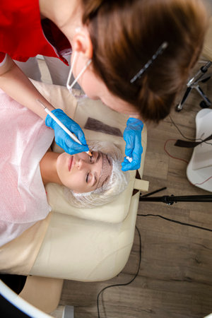 Closeup of Professional Female Beautician Tattooing Woman's Eyebrows Using Special Marking Equipment...