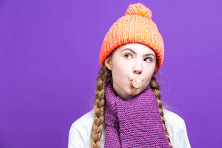 Concentrated Girl In Coral Knitted Seasonal Hat Posing With Chewing Gum Bubble In Front of Face Against Purple Seamless Background. Square Imageの写真素材