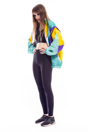 Full Length Portrait of Positive Winsome Caucasian Female Athlete Sportwoman in Fitness Skinny Outfit Wearing Colorful Windstopper Jacket While Posing in Training Over White. Vertical Imageの写真素材