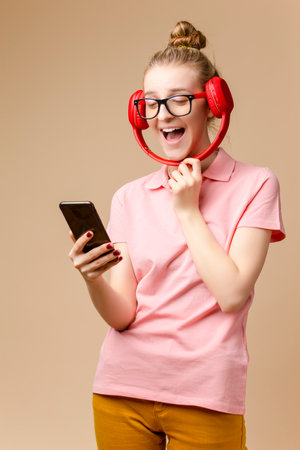 Winsome Happy Caucasian Girl In Casual Clothing Posing With Wireless Headphones With Cellphone And Exclaiming With Lifted Hand Against Beige. Vertical imageの写真素材