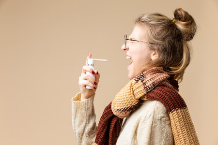 Portrait of Young Caucasian Female With Sore Throat Using Spray Inhaler for The Infection Protection and Disinfection Over Beige.Vertical Shotの写真素材