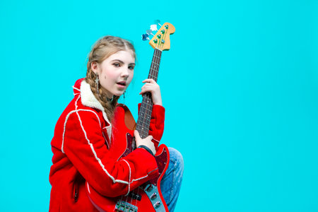 Relaxed Winsome Caucasian Guitar Player With Yellow Bass Guitar Posing In Fashionable Red Jacket Over Trendy Turquoise Background. Horizontal Shotの写真素材
