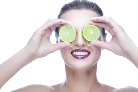 Portrait of Winsome Caucasian Woman with Perfect White Smile Posing With Green Lime Against Her Eyes On White Background. Horizontal Imageの写真素材