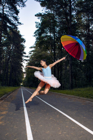 Young Caucasian Ballet Dancer in Light Orange Blue Tutu Posing in Dance Pose With Colorful Umbrella During Ballet Pas in Summer Forest With Lifted Hands Outdoor. Vertical image Orientationの写真素材