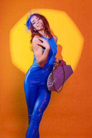 Sexy Caucasian Blond Woman in Blue Artistic Dance Suit Posing In Shawl Hat With Extravagant Hand-Bag Against Spot On Yellow Orange Background. Vertical Compositionの写真素材