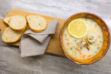 Brown Clay Bowl WIth Italian Lemon Chicken Orzo Soup With Freshly Baked White Bread Over Wooden...