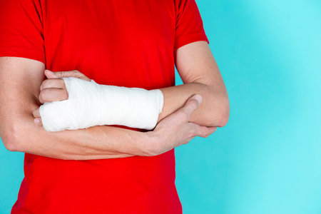 One Caucasian Man in Red Shirt with Broken Hand In Plaster and Bandaid Holding Hands Folded Isolated on Turquoise Background. Horizontal Imageの写真素材