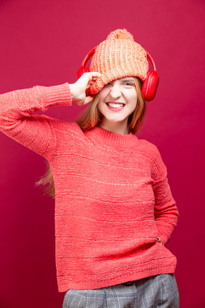 Happy Caucasian Girl In Coral knitted Sweater Posing With Wireless Headphones And Hat While Exclaiming With Lifted Hand Against Red. Vertical imageの写真素材