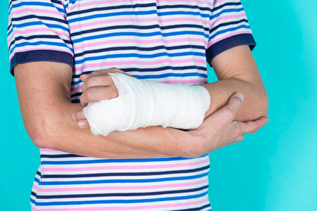 Man in Colorful Striped Shirt with Broken Hand In Plaster and Bandaid Holding Hands Folded Isolated on Turquoise Background. Horizontal Shotの写真素材