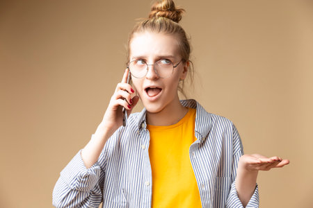 Portrait of Playful and Thoughful Beautiful Caucasian Blond in Striped Shirt and Glasses Using Cellphone Over Beige. Horizontal Imageの写真素材