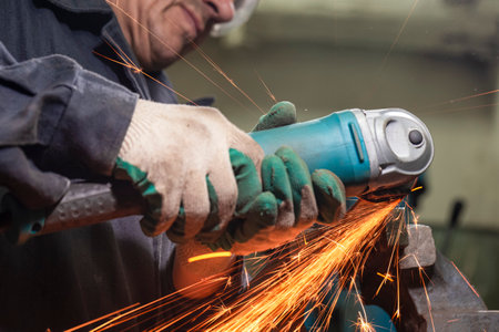 Hands of Male Fitter in Protective Gloves and Uniform With Gringer Tool For Metal Work With Bunch of Sparks in Workshop. Horizontal Orientation Imageの写真素材