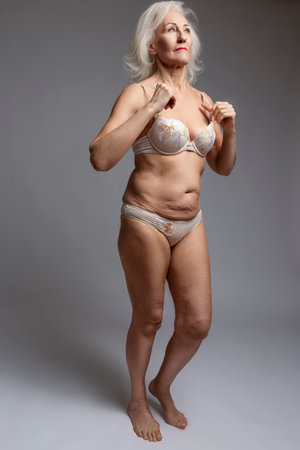 Full Length of Cute Calm Positive Mature Confident Caucasian Woman in White Lingerie With Hands Lifted Over White Background. Vertical Imageの写真素材