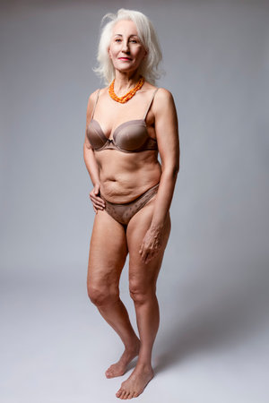 Full Length Natural Portrait of Mature Blond Caucasian Woman in Brown Lingerie Wearing Amber Necklace Posing Over Dark White Background.Vertical Image Compositionの写真素材