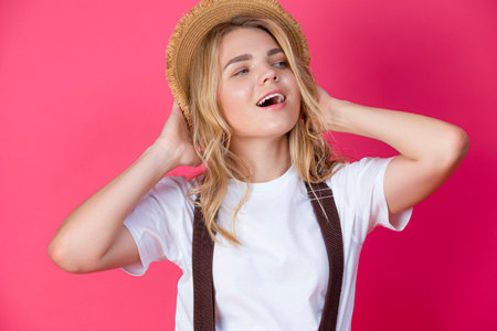 Cute Caucasian Blond Girl in Casual White Shirt With Suspenders Wearing Straw Hat Posing With Lifted hand Against Coral Background.Horizontal Shotの写真素材