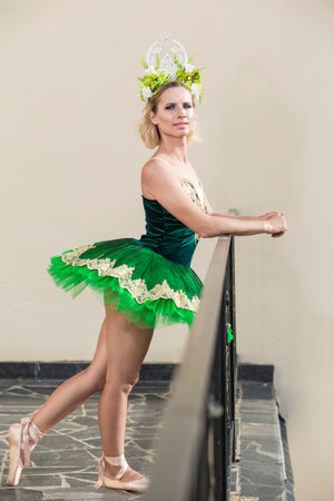 Smiling Calm Sensual Winsome Professional Caucasian Ballet Dancer in Green Tutu Dress Posing in dance Pose Wearing Flowery Crown Near Fence Outdoors. Vertical Imageの写真素材